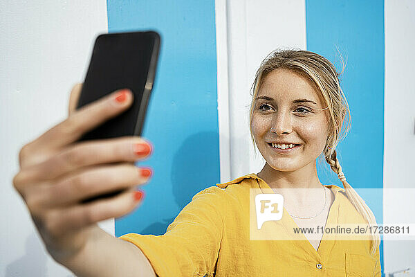 Lächelnde blonde Frau nimmt Selfie durch Handy von Wand