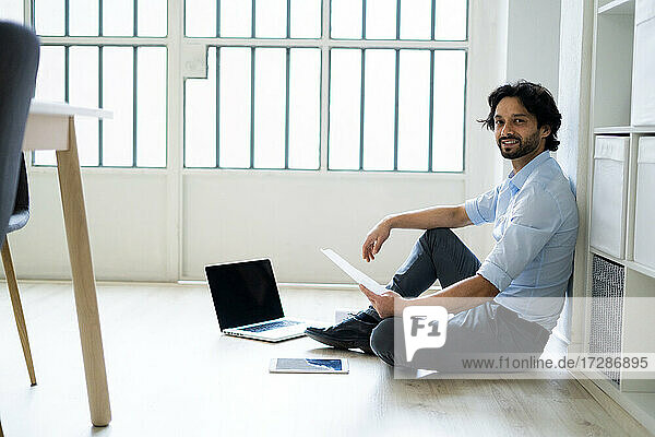Männlicher Unternehmer mit digitalem Tablet und Laptop auf dem Boden sitzend im Büro