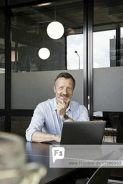 Männlicher Berufstätiger  der vor einem Laptop im Büro sitzt und lächelt