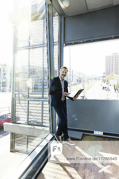 Männlicher Berufstätiger hält Laptop und lehnt sich an ein Glasfenster im Büro