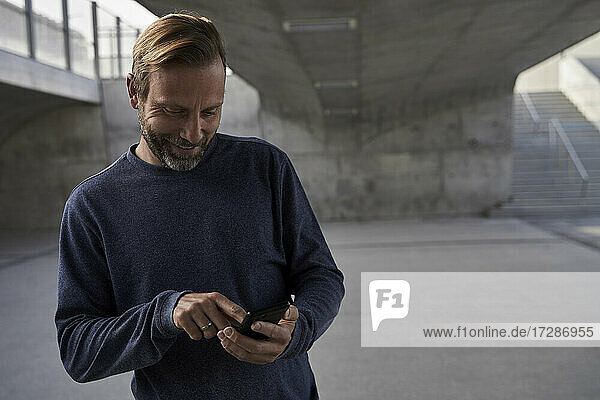 Lächelnder männlicher Berufstätiger  der unter einer Brücke stehend eine Textnachricht über sein Smartphone verschickt