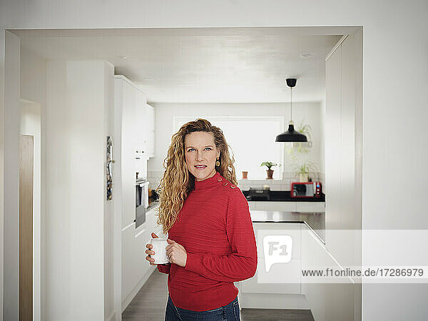 Lächelnde Frau mit Kaffeetasse in der Hand in der Küche stehend