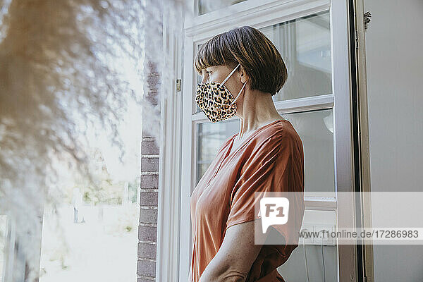 Frau mit Gesichtsmaske mit Tiermotiven schaut durch ein Fenster zu Hause
