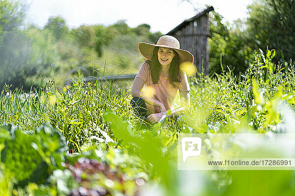 Lächelnde junge Frau mit Sonnenhut  die im Garten inmitten von Pflanzen hockt
