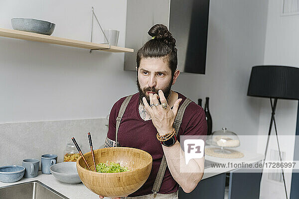 Bearded man tasting salad while standing in kitchen