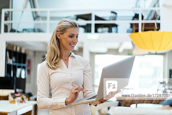 Lächelnde blonde Frau  die einen Laptop benutzt  während sie in einem kreativen Büro steht