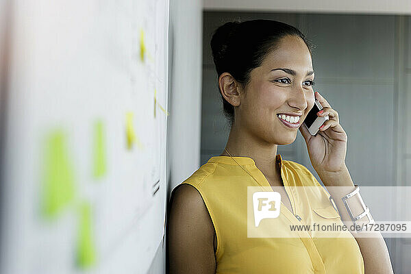 Lächelnde Geschäftsfrau  die wegschaut  während sie mit dem Handy im Büro telefoniert