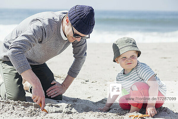 Father digging sand by son wearing hat looking away