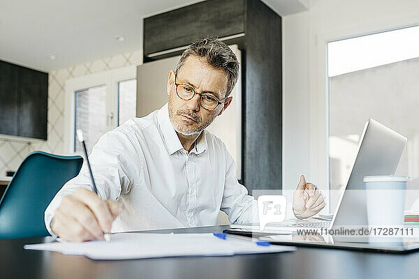 Älterer Geschäftsmann schreibt an einem Dokument  während er vor einem Laptop im Büro zu Hause sitzt
