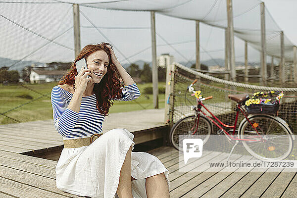 Lächelnde Frau mit dem Kopf in der Hand  die auf einem Pier sitzend mit ihrem Smartphone spricht