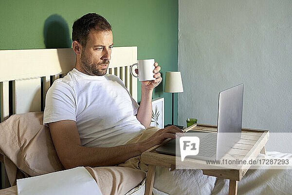 Mittlerer erwachsener Mann mit Kaffeetasse und Laptop  während er sich im Schlafzimmer ausruht