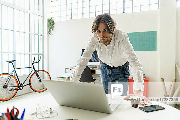 Businessman looking at laptop while leaning on desk in creative office