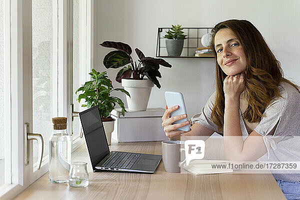 Schöne Frau mit Hand am Kinn hält Smartphone am Schreibtisch im Büro zu Hause