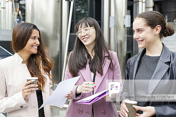 Lächelnde Geschäftsfrauen diskutieren im Stehen mit Kaffeetassen und Akten