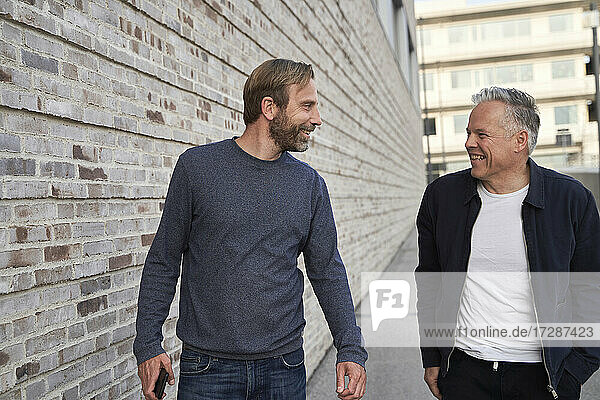 Happy businessmen looking at each other while walking by wall