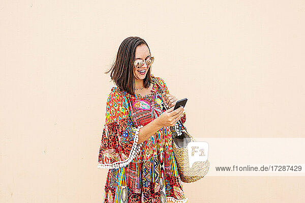 Fröhliche Frau mit Sonnenbrille  die ein Smartphone benutzt  während sie vor einer pfirsichfarbenen Wand steht