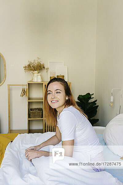 Smiling woman looking away while sitting on bed at home