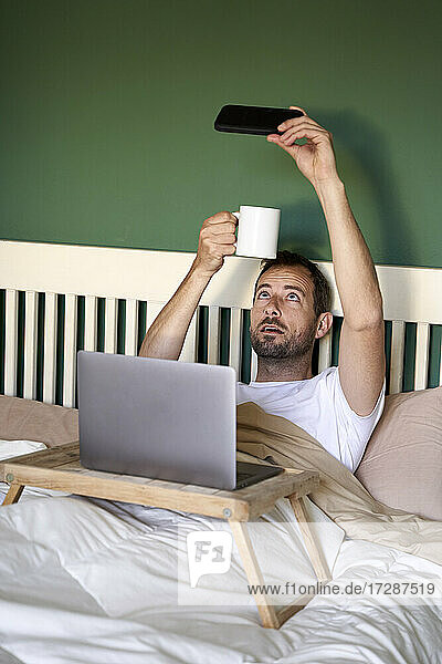 Mid erwachsenen Mann  der selfie mit Kaffeetasse durch Smartphone im Schlafzimmer