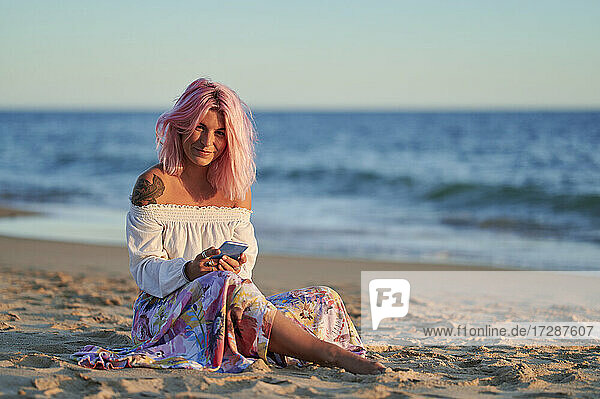 Frau mit rosafarbenen Haaren  die ihr Handy hält  während sie bei Sonnenuntergang am Strand sitzt