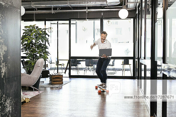 Lächelnde männliche Fachkraft mit Laptop fährt Skateboard im Büro