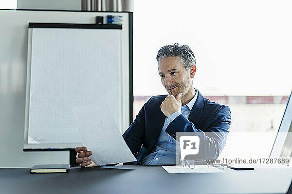 Mature businessman with hand on chin reading document sitting at desk in office