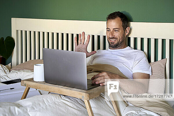 Smiling man waving hand to video call through laptop at home