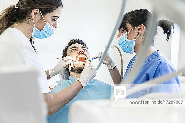 Weibliche Zahnärzte beim Bohren von Zähnen eines männlichen Patienten in einer Klinik während der Pandemie