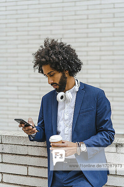 Männlicher Unternehmer mit Einwegbecher  der an der Wand lehnend ein Mobiltelefon benutzt