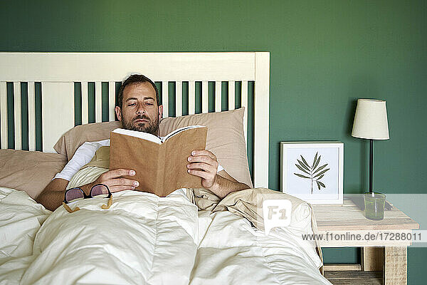 Mid adult man reading book while resting in bedroom