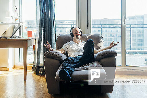 Smiling man wearing headphones gesturing while sitting on reclining chair in living room