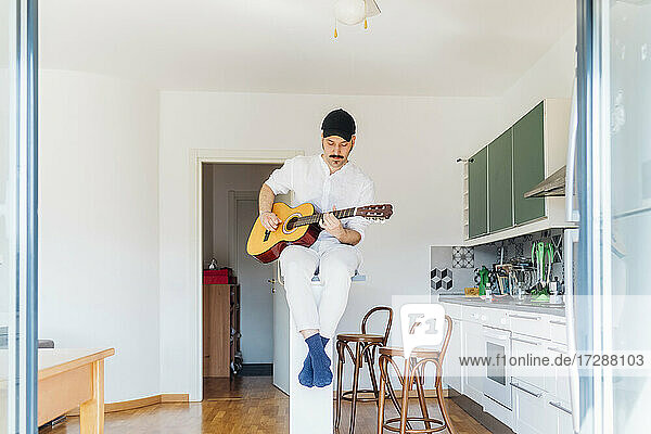 Männlicher Gitarrist mit Mütze spielt Gitarre in der Küche