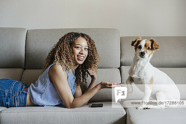 Lächelnde junge Frau  die dem Hund auf dem Sofa zu Hause einen Händedruck gibt
