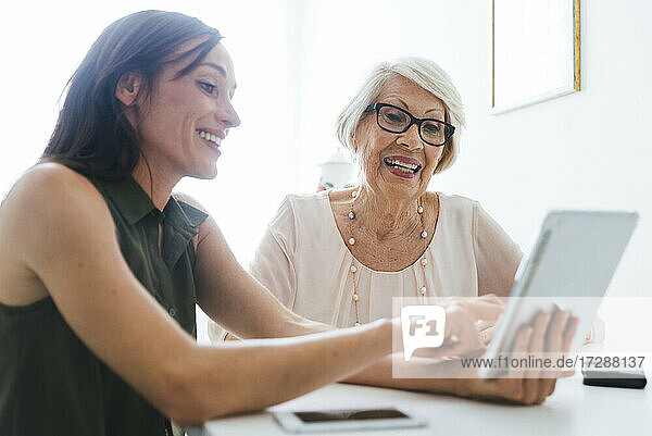 Lächelnde erwachsene Frau  die ihrer Großmutter ein digitales Tablet zeigt  während sie am Tisch sitzt