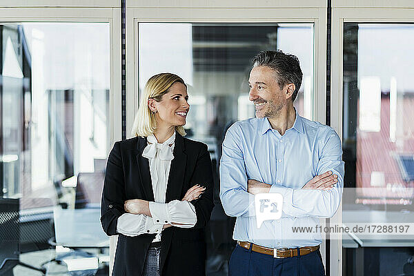 Lächelnde männliche und weibliche Fachkräfte mit verschränkten Armen  die sich gegenseitig ansehen  während sie an der Tür eines Büros stehen