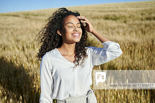 Lächelnde  lockig behaarte Frau mit geschlossenen Augen in einem Weizenfeld an einem sonnigen Tag