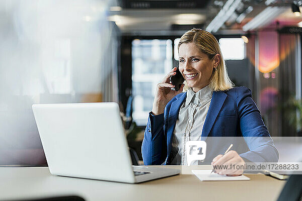 Lächelnde Unternehmerin  die mit einem Smartphone spricht  während sie mit einem Laptop am Schreibtisch sitzt