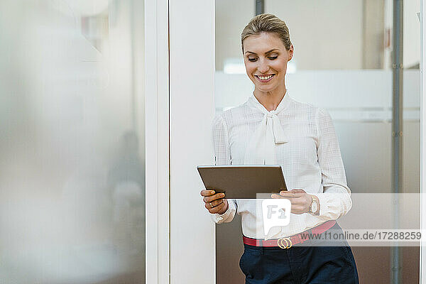 Lächelnde Unternehmerin  die ein digitales Tablet benutzt  während sie an der Tür im Büro lehnt