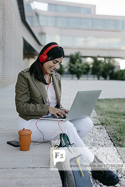 Lächelnde Geschäftsfrau mit Kopfhörern  die einen Laptop benutzt  während sie auf einem Fußweg sitzt