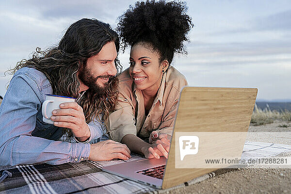 Mid adult couple smiling while lying by laptop on blanket during sunset
