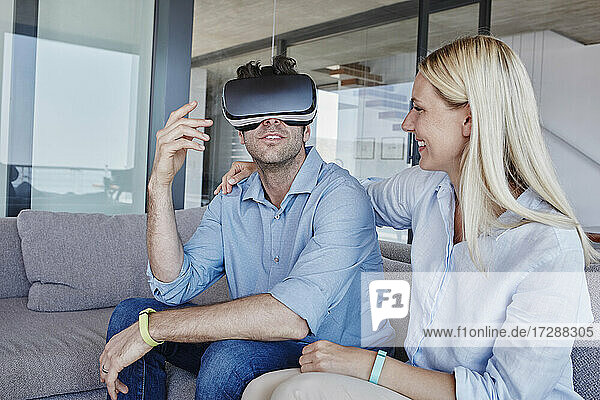 Fröhliche blonde Frau schaut auf einen Mann  der einen Virtual-Reality-Simulator im Wohnzimmer trägt