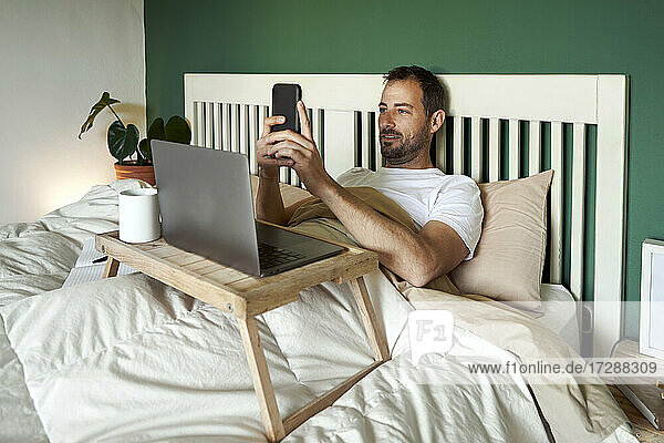 Man using mobile phone while lying with laptop on table at home