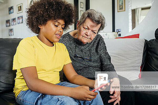 Ältere Frau sieht ihren Enkel an  der ein Smartphone benutzt  während sie auf dem Sofa im Wohnzimmer sitzt