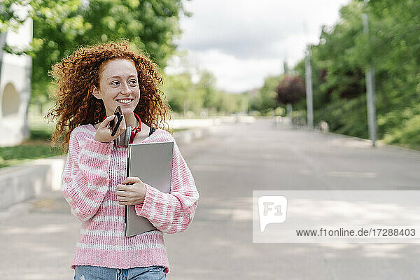 Lächelnde junge Frau mit lockigem Haar  die im Park über einen Lautsprecher mit ihrem Smartphone spricht und dabei wegschaut