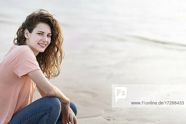 Lächelnde junge Frau  die am Strand im Sand hockt