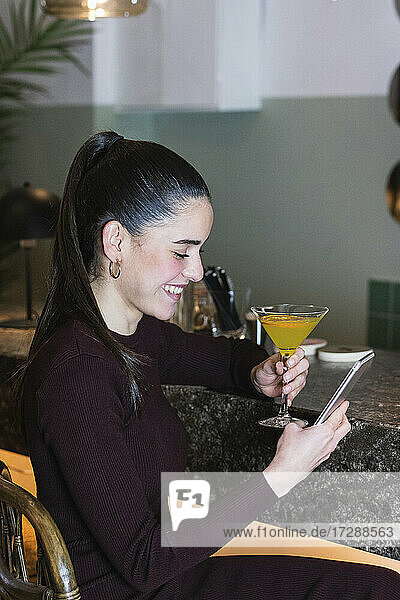 Lächelnde Frau mit Martini-Glas  die ein Mobiltelefon im Restaurant benutzt