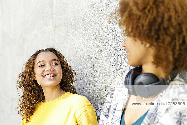Smiling young woman looking at female friend in front of wall