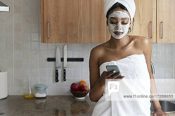Young woman wearing towel using mobile phone in kitchen