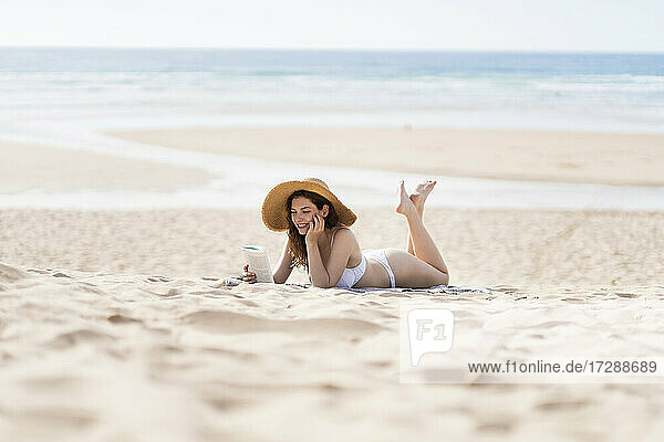 Junge Frau im Bikini liest ein Buch  während sie an einem sonnigen Tag am Strand im Sand liegt