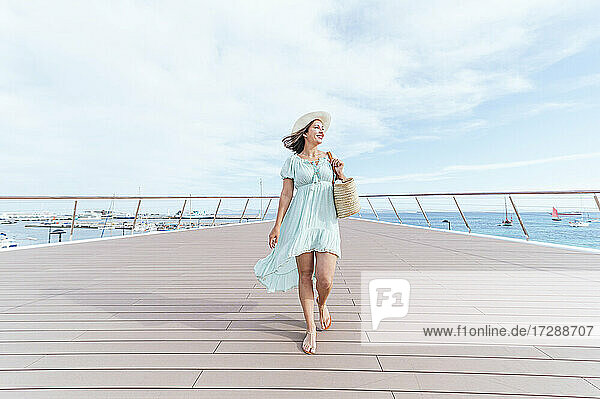 Lächelnde Frau mit Hut  die wegschaut  während sie auf dem Pier spazieren geht