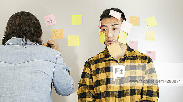 Verwirrter junger Geschäftsmann mit Haftnotizen im Gesicht neben einem männlichen Kollegen  der im Büro schreibt